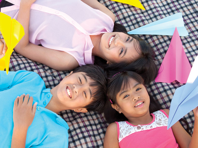 A boy and two girls laying on a blanket with paper planes.
